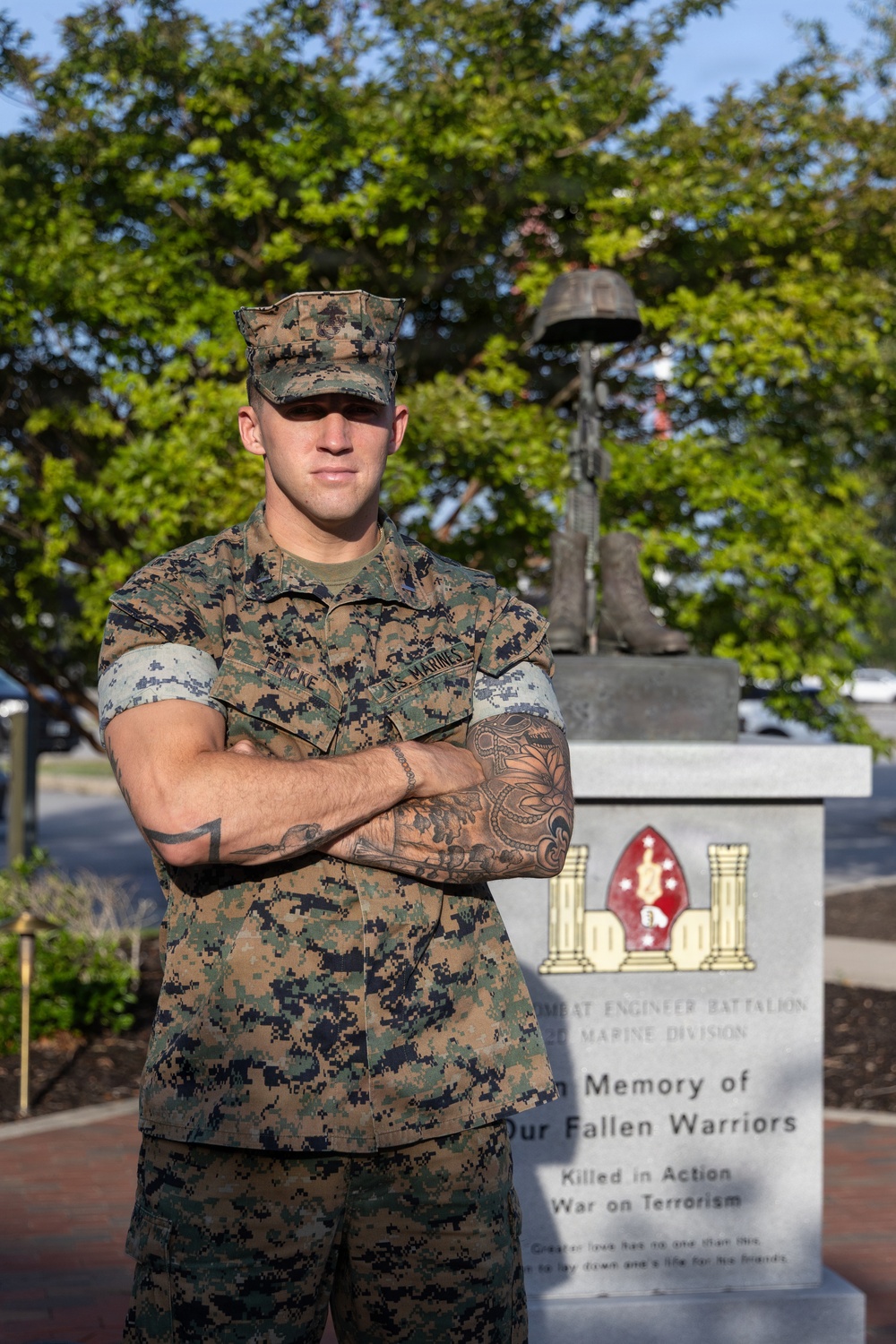 A U.S. Marine in MARPAT digital camouflage uniform stands with arms crossed in front of a military memorial monument. The memorial is dedicated to fallen warriors of a Combat Engineer Battalion, Marine Division, honoring those Killed in Action in the War on Terrorism. The setting appears to be an outdoor memorial area on a military installation, with green trees visible in the background.