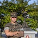 A U.S. Marine in MARPAT digital camouflage uniform stands with arms crossed in front of a military memorial monument. The memorial is dedicated to fallen warriors of a Combat Engineer Battalion, Marine Division, honoring those Killed in Action in the War on Terrorism. The setting appears to be an outdoor memorial area on a military installation, with green trees visible in the background.