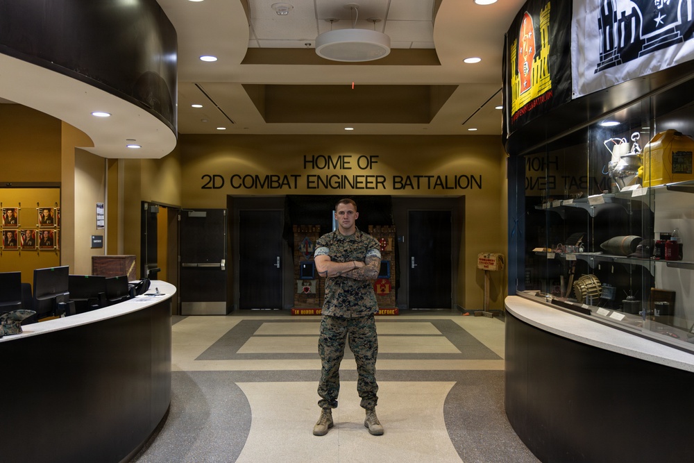 A U.S. Marine in digital camouflage (MARPAT) uniform stands with arms crossed in the lobby of the 2nd Combat Engineer Battalion headquarters. The lobby features a display case with military artifacts on the right, a reception desk on the left, and unit flags and memorabilia visible in the background. The signage on the wall reads 'HOME OF 2D COMBAT ENGINEER BATTALION.'