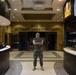 A U.S. Marine in digital camouflage (MARPAT) uniform stands with arms crossed in the lobby of the 2nd Combat Engineer Battalion headquarters. The lobby features a display case with military artifacts on the right, a reception desk on the left, and unit flags and memorabilia visible in the background. The signage on the wall reads 'HOME OF 2D COMBAT ENGINEER BATTALION.'