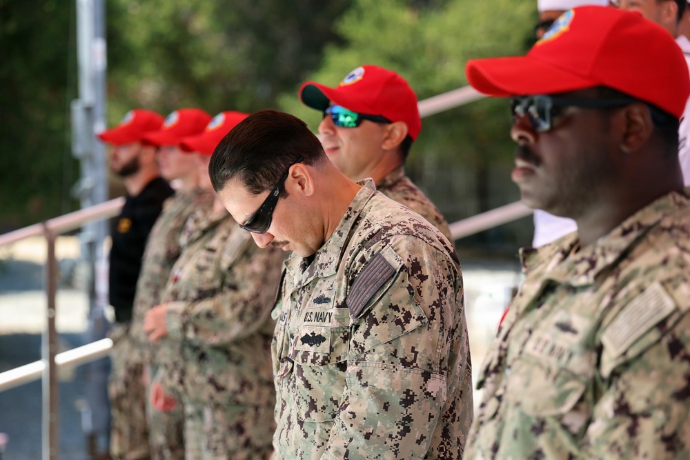 A group of U.S. Navy personnel in NWU Type III (woodland digital camouflage) uniforms are standing in formation, likely during a ceremony or official event. Several individuals are wearing distinctive red baseball caps, possibly indicating a specific unit, team, or event designation. The central figure has his head bowed, suggesting a moment of reflection, prayer, or formal observance.