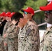 A group of U.S. Navy personnel in NWU Type III (woodland digital camouflage) uniforms are standing in formation, likely during a ceremony or official event. Several individuals are wearing distinctive red baseball caps, possibly indicating a specific unit, team, or event designation. The central figure has his head bowed, suggesting a moment of reflection, prayer, or formal observance.