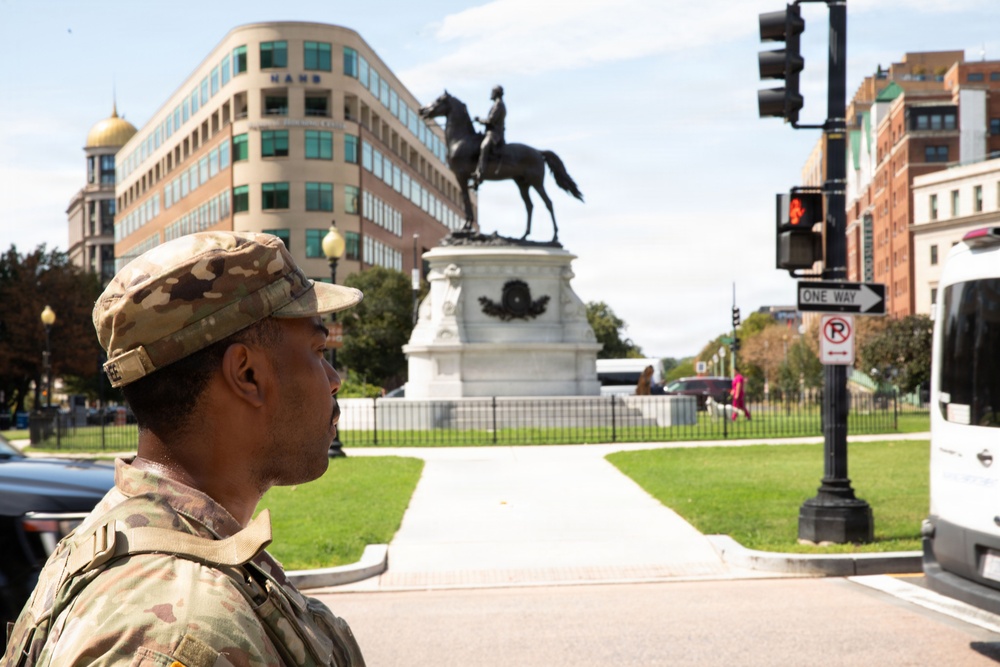 A U.S. Army soldier in MultiCam camouflage uniform stands at a street intersection in what appears to be Washington, D.C. In the background, a prominent equestrian statue on a white marble pedestal is visible within a fenced park area. Urban buildings, traffic signals, and street signs are visible in the surrounding environment.