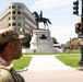 A U.S. Army soldier in MultiCam camouflage uniform stands at a street intersection in what appears to be Washington, D.C. In the background, a prominent equestrian statue on a white marble pedestal is visible within a fenced park area. Urban buildings, traffic signals, and street signs are visible in the surrounding environment.