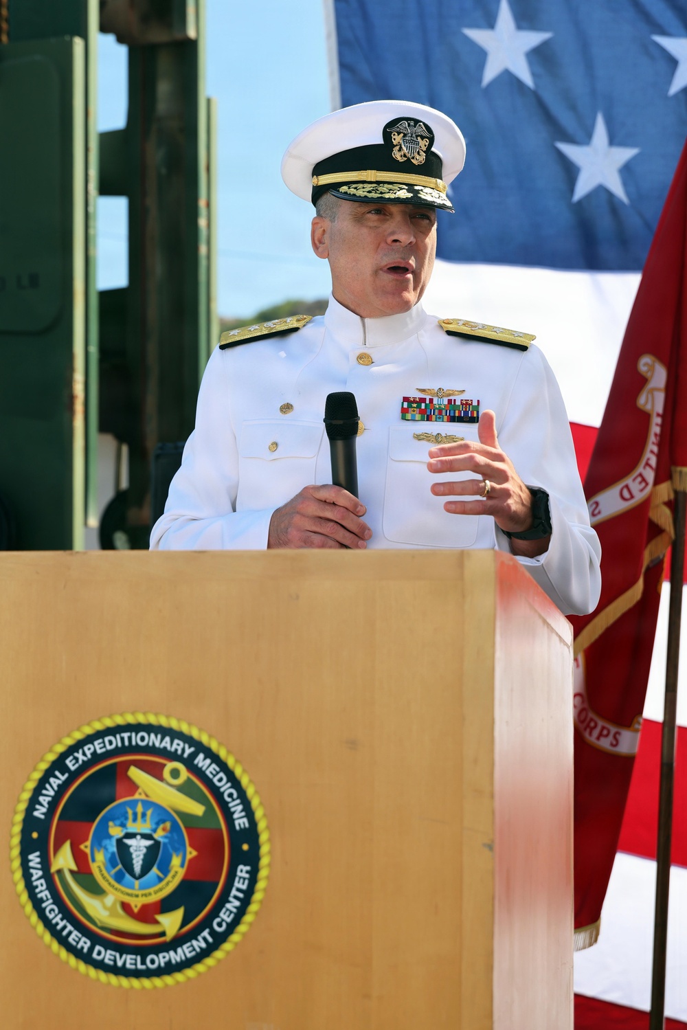 A senior U.S. Navy flag officer in white dress uniform is speaking at a podium bearing the seal of the Naval Expeditionary Medicine Warfighter Development Center. He is holding a microphone and gesturing with his right hand, suggesting an active address or speech. An American flag and what appears to be a Marine Corps flag are visible in the background.