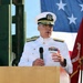A senior U.S. Navy flag officer in white dress uniform is speaking at a podium bearing the seal of the Naval Expeditionary Medicine Warfighter Development Center. He is holding a microphone and gesturing with his right hand, suggesting an active address or speech. An American flag and what appears to be a Marine Corps flag are visible in the background.