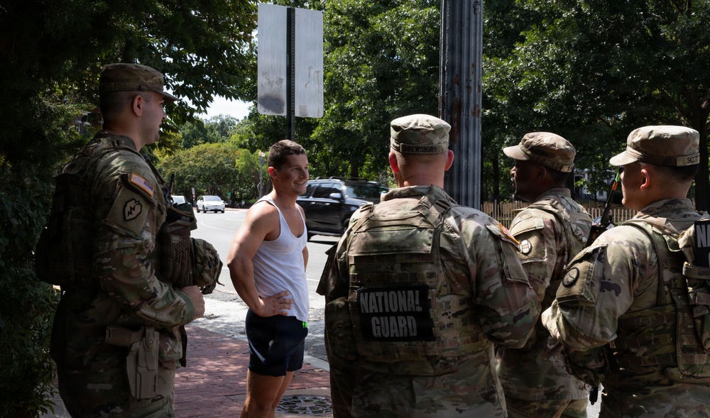Four National Guard soldiers in camouflage uniforms are conversing with a civilian man wearing a white tank top and blue shorts on a brick sidewalk. The interaction appears casual and friendly, taking place in an urban setting with trees and street traffic visible in the background. The soldiers are equipped with tactical gear and body armor marked with 'NATIONAL GUARD' insignia.