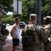 Four National Guard soldiers in camouflage uniforms are conversing with a civilian man wearing a white tank top and blue shorts on a brick sidewalk. The interaction appears casual and friendly, taking place in an urban setting with trees and street traffic visible in the background. The soldiers are equipped with tactical gear and body armor marked with 'NATIONAL GUARD' insignia.