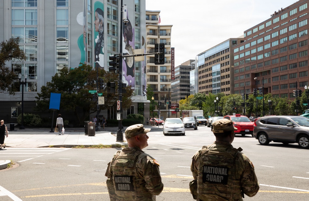 Two National Guard soldiers in camouflage uniforms stand at a busy urban intersection, viewed from behind. The scene appears to be in Washington, D.C., identifiable by the street signs and a visible 'Residence Inn' hotel sign. A large colorful mural depicting figures is painted on the side of a modern building in the background.