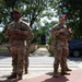 Two U.S. Army soldiers in OCP (Operational Camouflage Pattern) uniforms are standing at a crosswalk intersection, appearing to be on guard or security duty. A street sign reading 'Logan' is visible in the background, along with a fenced park area containing a statue or monument. The scene suggests a military security presence in an urban area.
