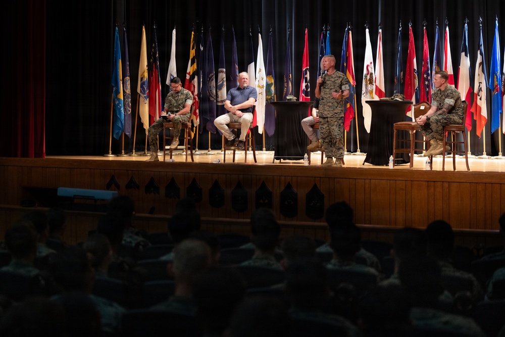 A military panel discussion or briefing is taking place on a stage in an auditorium. Four individuals — three in military uniforms and one in civilian attire — are seated or standing on stage, with one uniformed speaker holding a microphone. A large audience of uniformed personnel fills the seating area in the foreground.