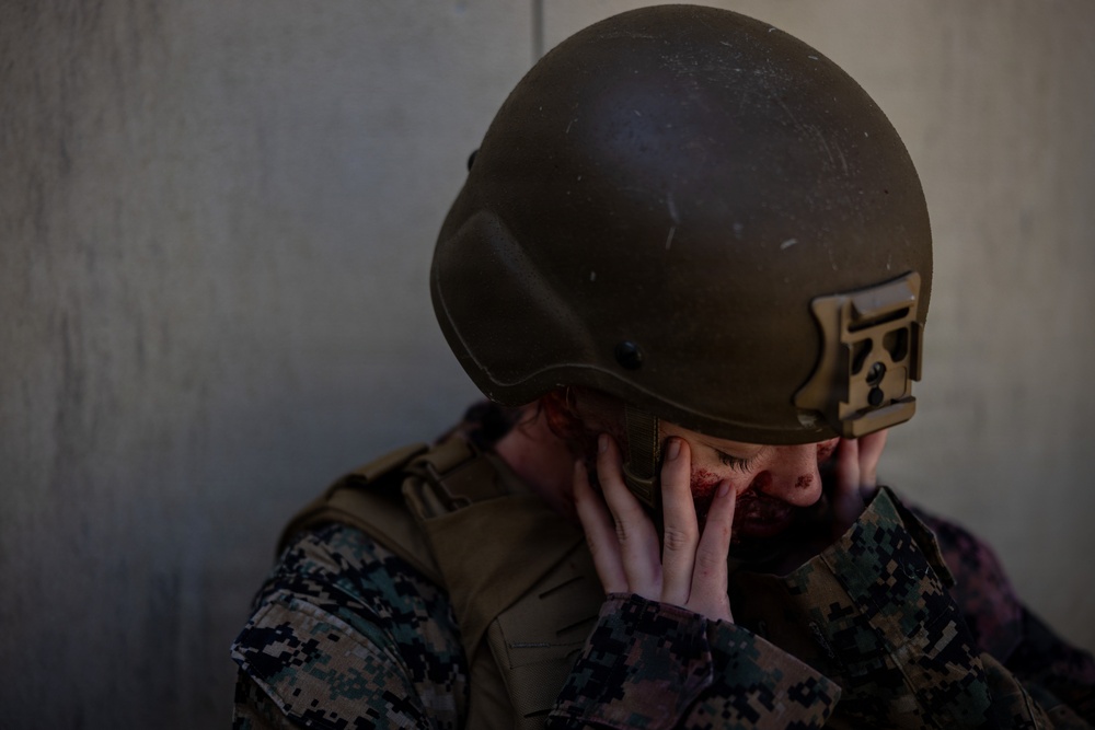 A military service member wearing digital camouflage uniform and olive drab tactical helmet is shown with their head bowed and hands covering their face, which shows visible redness or simulated blood. The individual appears to be in distress or participating in a combat casualty training exercise. The setting is against a plain concrete or stucco wall.