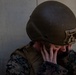 A military service member wearing digital camouflage uniform and olive drab tactical helmet is shown with their head bowed and hands covering their face, which shows visible redness or simulated blood. The individual appears to be in distress or participating in a combat casualty training exercise. The setting is against a plain concrete or stucco wall.
