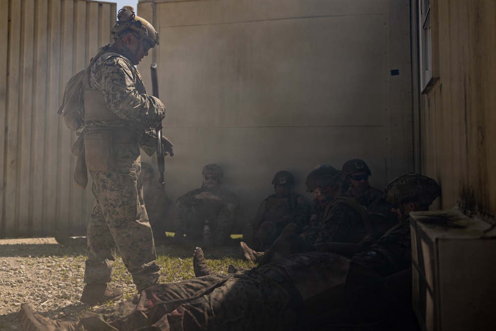 The image depicts military personnel in a tactical scenario, likely a training exercise, near modular building structures. Smoke is visible in the background, and one standing soldier in digital camouflage appears to be overseeing or directing several seated/crouching personnel. A figure is visible lying on the ground in the foreground.