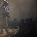 The image depicts military personnel in a tactical scenario, likely a training exercise, near modular building structures. Smoke is visible in the background, and one standing soldier in digital camouflage appears to be overseeing or directing several seated/crouching personnel. A figure is visible lying on the ground in the foreground.