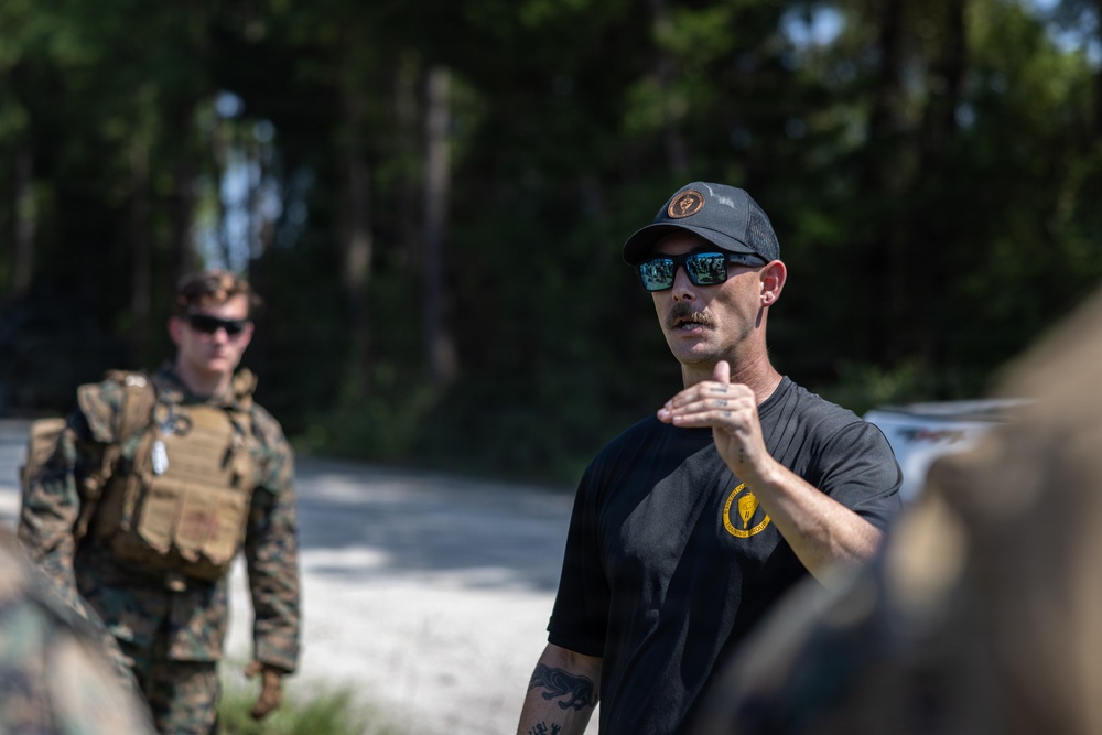 A civilian instructor or contractor in a black t-shirt and cap appears to be briefing or instructing a group in an outdoor wooded area. In the background, a uniformed military personnel wearing MARPAT camouflage and a plate carrier vest is visible. The setting suggests a military training exercise or instruction session.