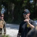 A civilian instructor or contractor in a black t-shirt and cap appears to be briefing or instructing a group in an outdoor wooded area. In the background, a uniformed military personnel wearing MARPAT camouflage and a plate carrier vest is visible. The setting suggests a military training exercise or instruction session.