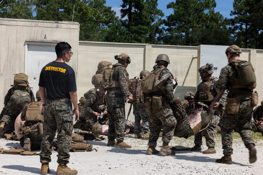 A military training exercise is underway at what appears to be a combat casualty care training facility. An instructor in a black 'INSTRUCTOR' shirt observes as multiple Marines in full combat gear practice tactical combat casualty care (TCCC) on simulated casualties. The scene takes place against a concrete training structure with pine trees visible in the background.