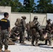 A military training exercise is underway at what appears to be a combat casualty care training facility. An instructor in a black 'INSTRUCTOR' shirt observes as multiple Marines in full combat gear practice tactical combat casualty care (TCCC) on simulated casualties. The scene takes place against a concrete training structure with pine trees visible in the background.