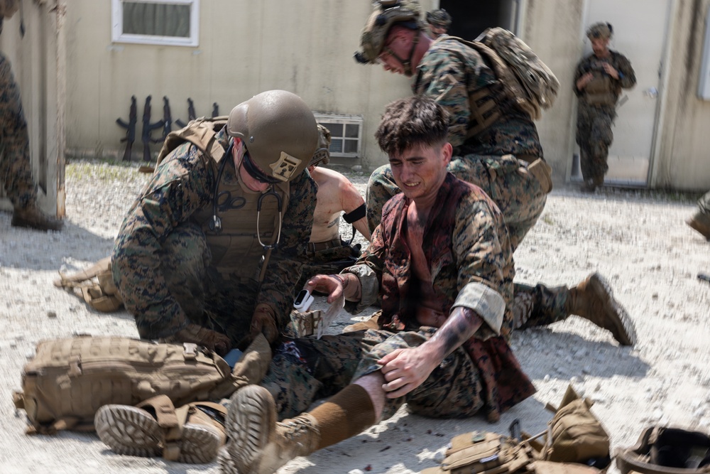 The image depicts what appears to be a military combat casualty care training exercise. Personnel in MARPAT (Marine pattern) camouflage uniforms are providing simulated medical treatment to a role-player acting as a casualty. The scene includes simulated blood/wounds and medical equipment consistent with tactical combat casualty care (TCCC) training.