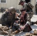The image depicts what appears to be a military combat casualty care training exercise. Personnel in MARPAT (Marine pattern) camouflage uniforms are providing simulated medical treatment to a role-player acting as a casualty. The scene includes simulated blood/wounds and medical equipment consistent with tactical combat casualty care (TCCC) training.