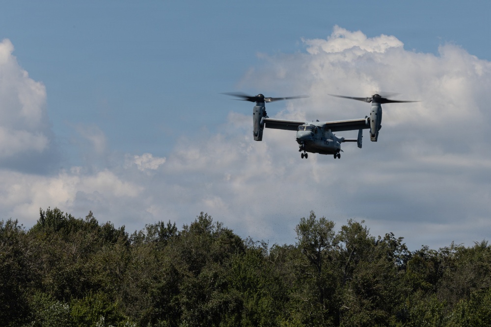 A military tiltrotor aircraft, identifiable as a Bell Boeing V-22 Osprey, is captured in low-altitude flight above a treeline. The aircraft appears to be in helicopter mode with rotors tilted upward, suggesting it is either landing or hovering. The image was taken in daylight under partly cloudy skies.