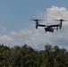 A military tiltrotor aircraft, identifiable as a Bell Boeing V-22 Osprey, is captured in low-altitude flight above a treeline. The aircraft appears to be in helicopter mode with rotors tilted upward, suggesting it is either landing or hovering. The image was taken in daylight under partly cloudy skies.