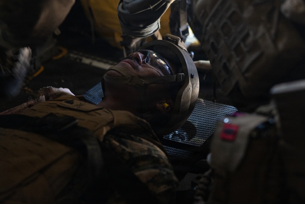 A military service member in combat uniform and helmet is lying on a litter/stretcher, appearing to be receiving medical attention. Multiple personnel are visible around the casualty in what appears to be a low-light environment, possibly inside a military aircraft or vehicle. The scene is consistent with a tactical casualty care or medical evacuation (MEDEVAC) scenario.