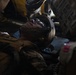 A military service member in combat uniform and helmet is lying on a litter/stretcher, appearing to be receiving medical attention. Multiple personnel are visible around the casualty in what appears to be a low-light environment, possibly inside a military aircraft or vehicle. The scene is consistent with a tactical casualty care or medical evacuation (MEDEVAC) scenario.