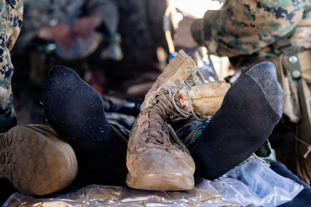 The image shows a close-up of a casualty's legs and feet during what appears to be a military medical training exercise or field treatment scenario. One boot remains on the foot while the other foot is in a black sock, suggesting a boot was removed for medical assessment. Personnel in MARPAT (Marine pattern) camouflage uniforms are visible in the background attending to the casualty.