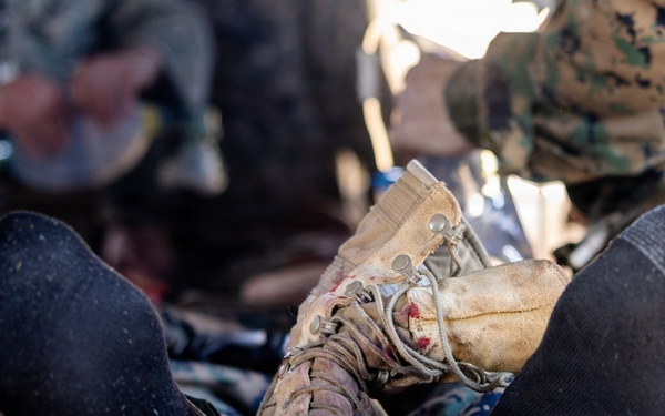 The image shows a close-up of a casualty's legs and feet during what appears to be a military medical training exercise or field treatment scenario. One boot remains on the foot while the other foot is in a black sock, suggesting a boot was removed for medical assessment. Personnel in MARPAT (Marine pattern) camouflage uniforms are visible in the background attending to the casualty.