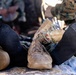 The image shows a close-up of a casualty's legs and feet during what appears to be a military medical training exercise or field treatment scenario. One boot remains on the foot while the other foot is in a black sock, suggesting a boot was removed for medical assessment. Personnel in MARPAT (Marine pattern) camouflage uniforms are visible in the background attending to the casualty.