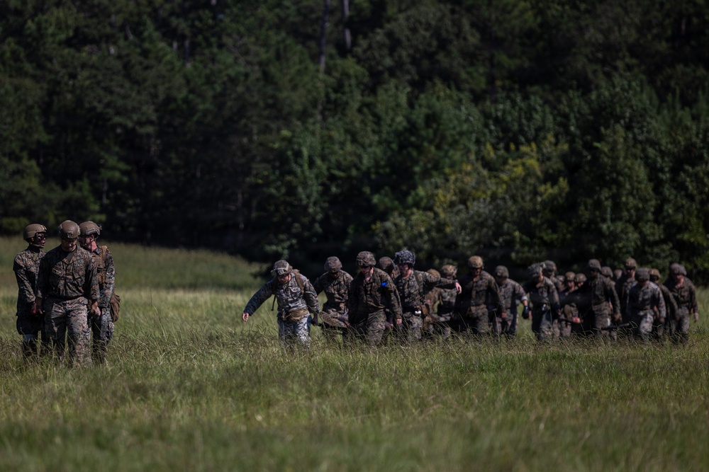 A column of military personnel in camouflage uniforms and combat helmets marches across an open grassy field. The group appears to be on a training exercise, moving in a loose formation from right to left across the frame. A dense tree line of deciduous and coniferous trees forms the background.