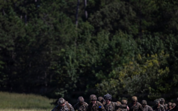 A column of military personnel in camouflage uniforms and combat helmets marches across an open grassy field. The group appears to be on a training exercise, moving in a loose formation from right to left across the frame. A dense tree line of deciduous and coniferous trees forms the background.