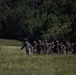 A column of military personnel in camouflage uniforms and combat helmets marches across an open grassy field. The group appears to be on a training exercise, moving in a loose formation from right to left across the frame. A dense tree line of deciduous and coniferous trees forms the background.