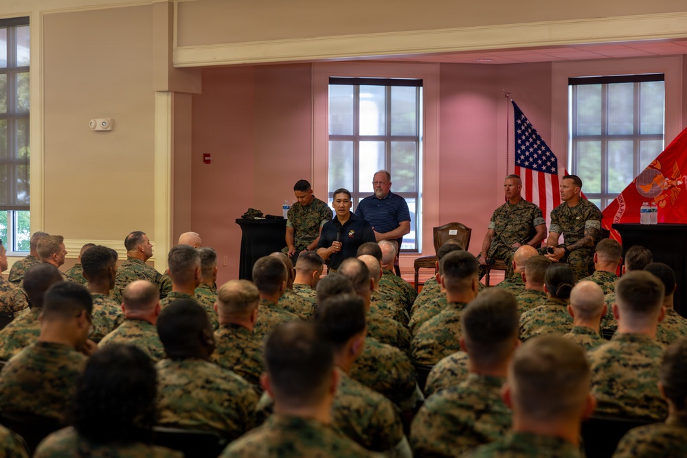 A large group of U.S. Marines in woodland camouflage uniforms are seated in an auditorium or meeting hall, attending a briefing or presentation. At the front of the room, several individuals including uniformed Marines and two civilians in polo shirts appear to be conducting or participating in a panel discussion or address. An American flag and what appears to be a Marine Corps flag are visible in the background.
