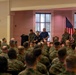 A large group of U.S. Marines in woodland camouflage uniforms are seated in an auditorium or meeting hall, attending a briefing or presentation. At the front of the room, several individuals including uniformed Marines and two civilians in polo shirts appear to be conducting or participating in a panel discussion or address. An American flag and what appears to be a Marine Corps flag are visible in the background.