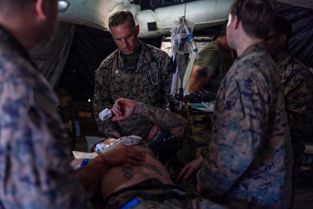Military medical personnel in MARPAT camouflage uniforms are providing medical treatment to a patient lying on a stretcher inside what appears to be a field medical tent. A central figure wearing a stethoscope, identifiable as a medical officer, is actively attending to the patient. The scene depicts a tactical combat casualty care or field medical training/operational scenario.