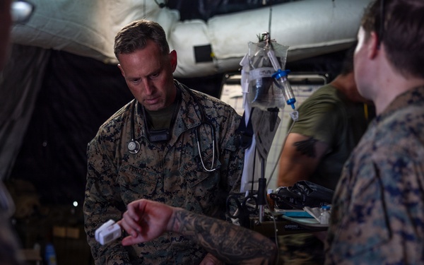Military medical personnel in MARPAT camouflage uniforms are providing medical treatment to a patient lying on a stretcher inside what appears to be a field medical tent. A central figure wearing a stethoscope, identifiable as a medical officer, is actively attending to the patient. The scene depicts a tactical combat casualty care or field medical training/operational scenario.