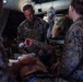 Military medical personnel in MARPAT camouflage uniforms are providing medical treatment to a patient lying on a stretcher inside what appears to be a field medical tent. A central figure wearing a stethoscope, identifiable as a medical officer, is actively attending to the patient. The scene depicts a tactical combat casualty care or field medical training/operational scenario.