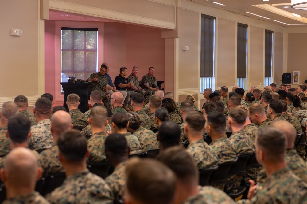 A large group of military personnel in camouflage uniforms are seated in a conference or event hall, facing a small panel of four individuals at the front of the room. The panel appears to consist of three uniformed military members and one civilian in a dark shirt, suggesting a briefing, town hall, or panel discussion format. The setting appears to be an indoor military facility or base conference center.