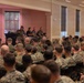 A large group of military personnel in camouflage uniforms are seated in a conference or event hall, facing a small panel of four individuals at the front of the room. The panel appears to consist of three uniformed military members and one civilian in a dark shirt, suggesting a briefing, town hall, or panel discussion format. The setting appears to be an indoor military facility or base conference center.