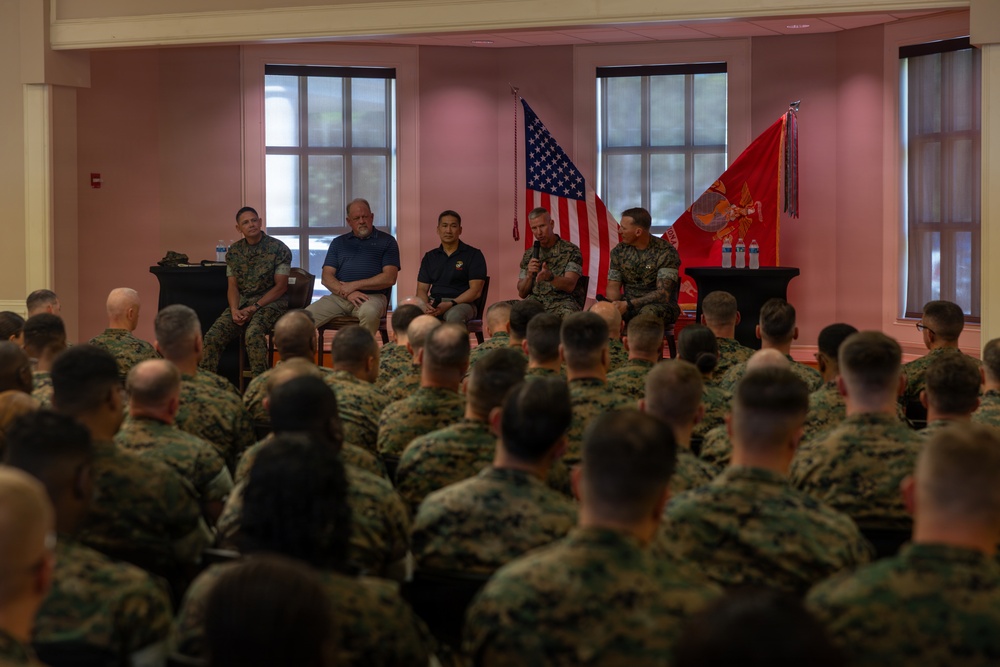 A U.S. Marine Corps briefing or town hall event is taking place in an indoor venue. A panel of five individuals — including uniformed Marines and two civilians in civilian attire — are seated on a stage addressing a large audience of Marines in camouflage uniforms. The American flag and the U.S. Marine Corps flag are prominently displayed on the stage behind the panel.