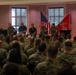 A U.S. Marine Corps briefing or town hall event is taking place in an indoor venue. A panel of five individuals — including uniformed Marines and two civilians in civilian attire — are seated on a stage addressing a large audience of Marines in camouflage uniforms. The American flag and the U.S. Marine Corps flag are prominently displayed on the stage behind the panel.