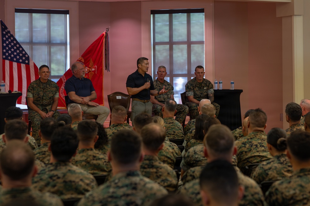 A formal military briefing or town hall is taking place in an indoor venue, with a panel of individuals seated at the front addressing a large audience of uniformed U.S. Marines. One civilian and several Marines in camouflage uniforms are seated on the panel, with one individual standing and speaking into a microphone. The U.S. flag and a red Marine Corps unit guidon are displayed prominently in the background.