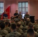 A formal military briefing or town hall is taking place in an indoor venue, with a panel of individuals seated at the front addressing a large audience of uniformed U.S. Marines. One civilian and several Marines in camouflage uniforms are seated on the panel, with one individual standing and speaking into a microphone. The U.S. flag and a red Marine Corps unit guidon are displayed prominently in the background.