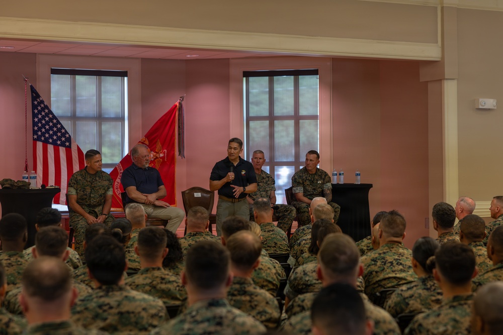 A military briefing or town hall event is taking place in an indoor venue, likely on a U.S. Marine Corps installation. A speaker in a black polo shirt and khaki pants is addressing a large audience of Marines in woodland MARPAT camouflage uniforms. Several individuals, including uniformed Marines and one civilian in a blue polo shirt, are seated on a panel behind the speaker.