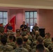 A military briefing or town hall event is taking place in an indoor venue, likely on a U.S. Marine Corps installation. A speaker in a black polo shirt and khaki pants is addressing a large audience of Marines in woodland MARPAT camouflage uniforms. Several individuals, including uniformed Marines and one civilian in a blue polo shirt, are seated on a panel behind the speaker.