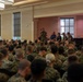 A large indoor military briefing or assembly is taking place in a formal hall, with hundreds of uniformed personnel seated and facing a stage area. One individual in military camouflage uniform is standing and speaking into a microphone, while several others are seated on the stage behind them. An American flag is prominently displayed on the right side of the stage.