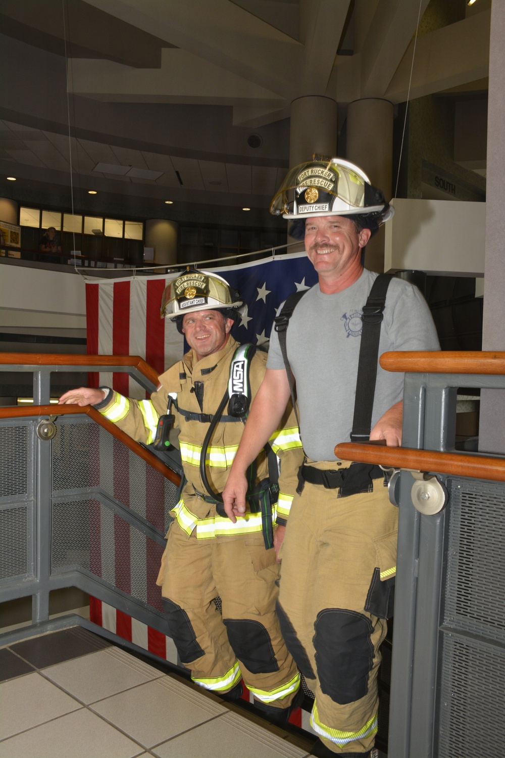 Two firefighters from Fort Rucker Fire &amp; Rescue are posing on a staircase inside a building, smiling at the camera. The firefighter on the left wears full turnout gear with an MSA SCBA unit and a helmet labeled 'Assistant Chief,' while the one on the right wears partial gear with a white helmet labeled 'Deputy Chief.' An American flag is displayed on the wall behind them.